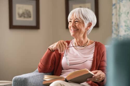 Cheerful senior woman holding book and eyeglasses thinking while relaxing at home. Happy elderly woman reading book at home sitting on couch. Beautiful old teacher takes a break from reading while looking through the window with a big grin.